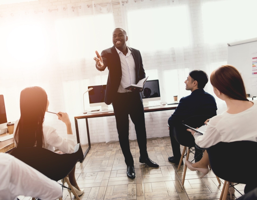 A black man talks to employees of a business office at a seminar. He has a good mood, he smiles at all.